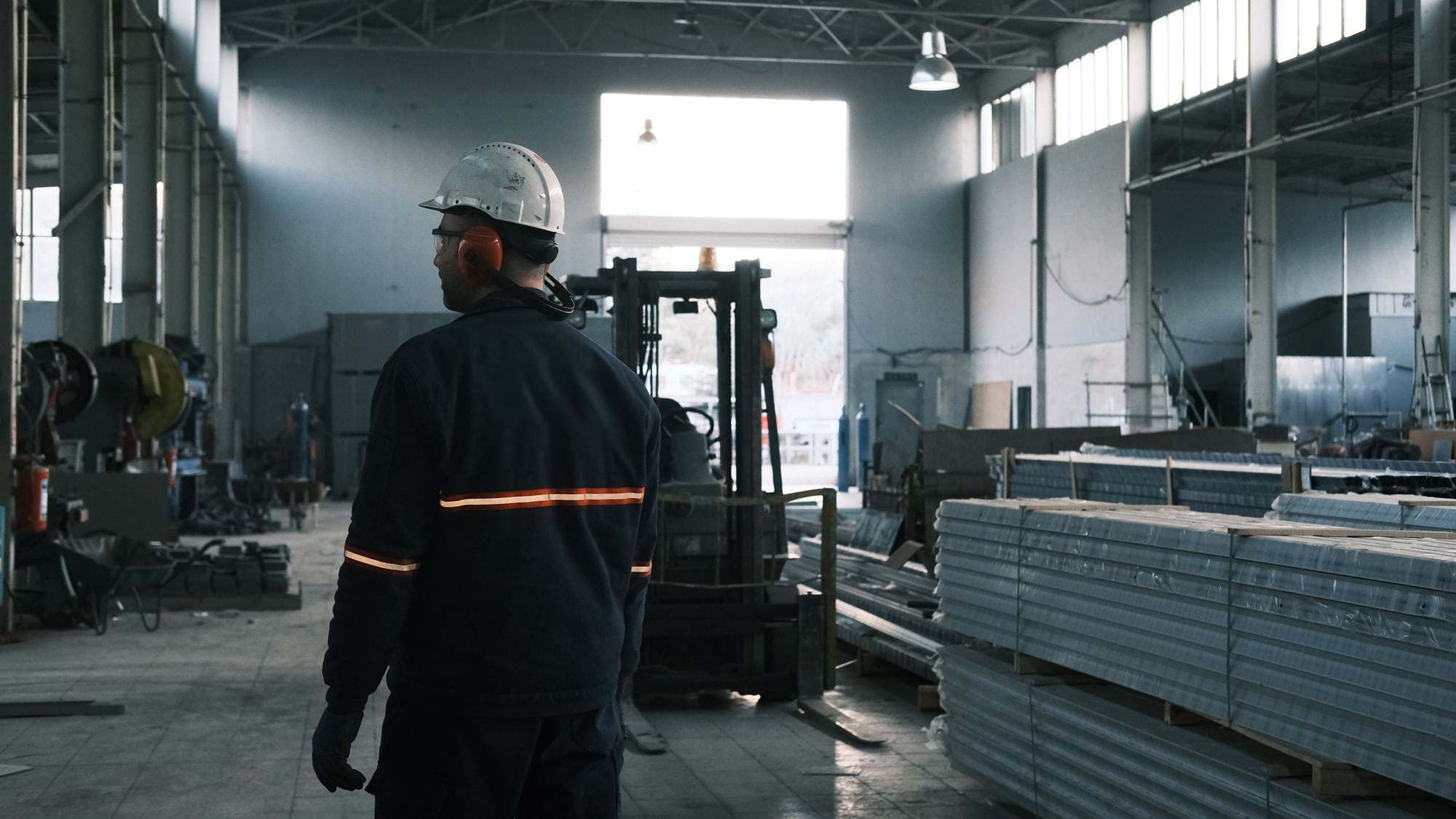 Mitarbeiter mit Schutzhelm und Gehörschutz in einer Produktionshalle, im Hintergrund ein Stapler